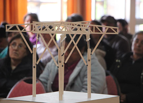 Maqueta de una estructura de madera en primer plano. En el fondo, desenfocado varias personas sentadas 